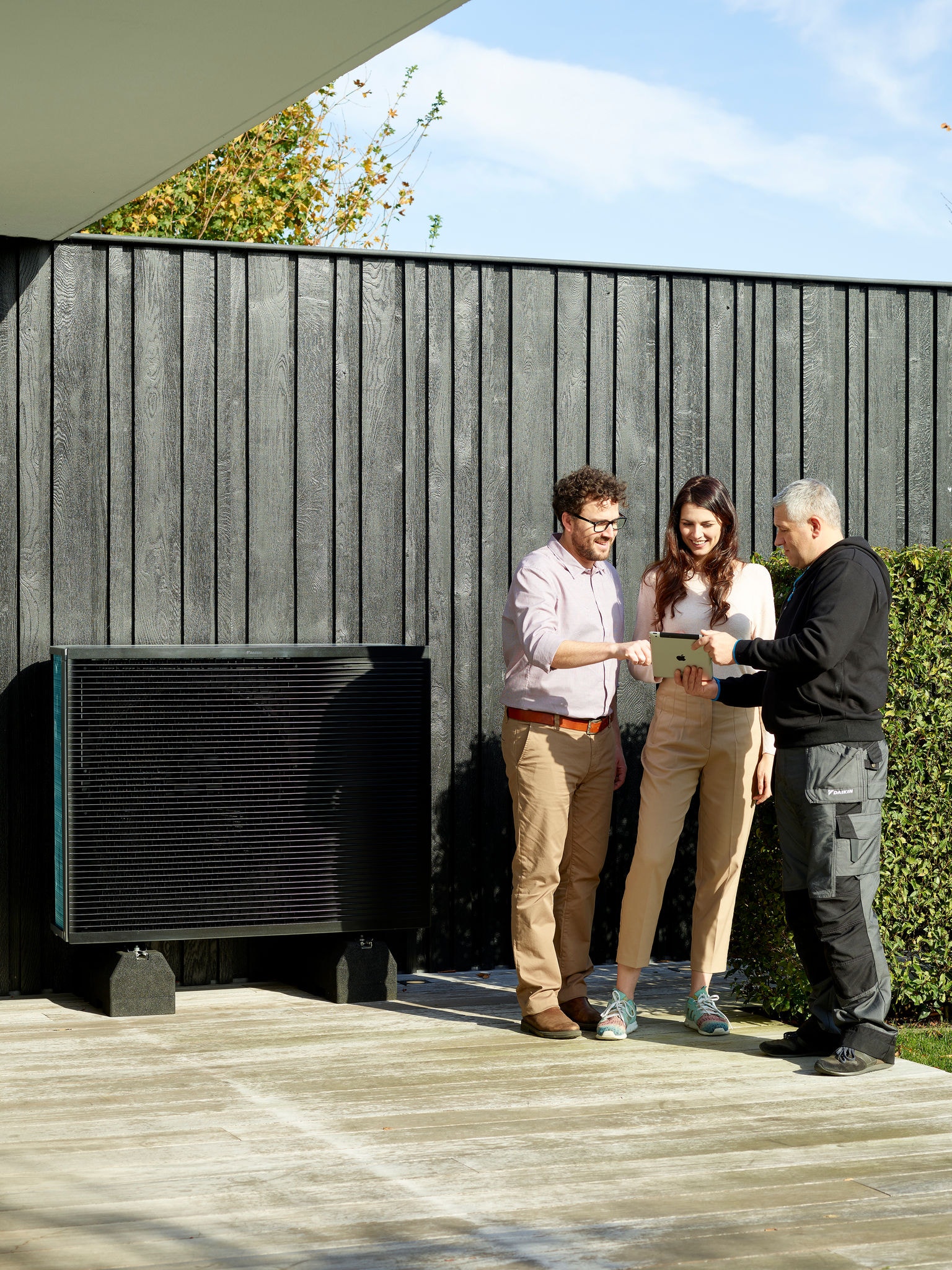 Couple talking to installer while looking on a tablet and standing next to the outside unit of a Daikin Altherma